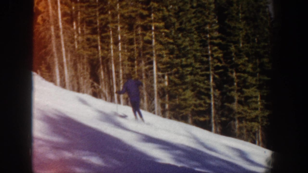 A small figure in dark blue skis farther down the Aspen snow slope, framed by tall evergreens and birch-like trunks.