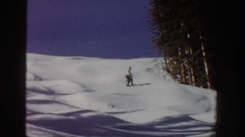 A small figure skis across an open snowy area below trees in Aspen, Colorado archival 8mm footage.