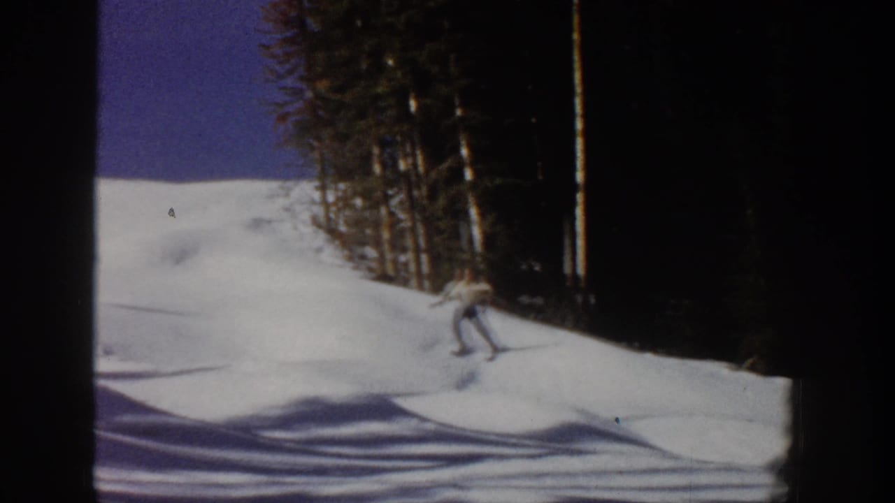 1961 Aspen home-movie frame showing a skier moving downhill on a snow-covered run bordered by dark evergreen trees.