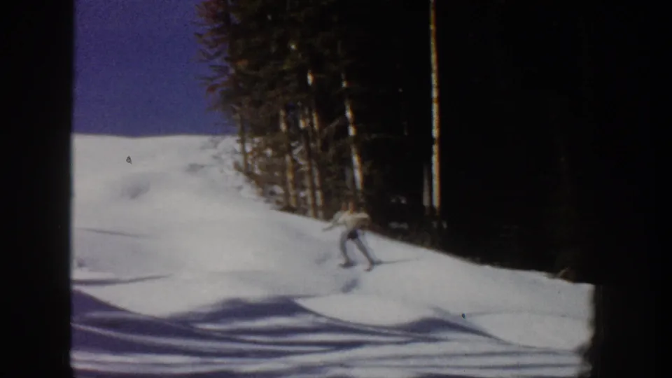 1961 Aspen home-movie frame showing a skier moving downhill on a snow-covered run bordered by dark evergreen trees.