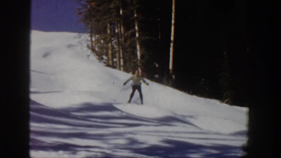 1961 Aspen frame with a skier in a light jacket and dark pants on a snow-covered slope lined by trees.