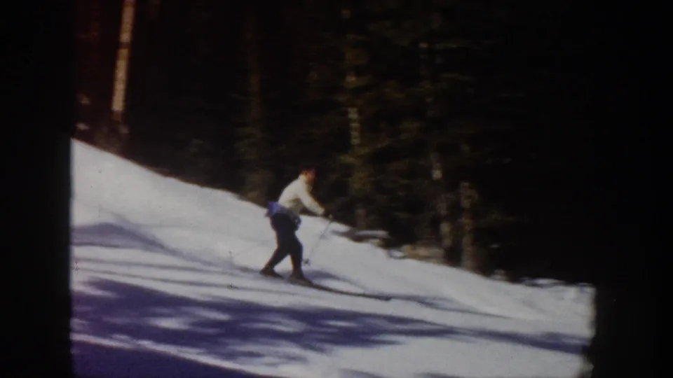 1961 Aspen home-movie still showing a skier moving across a snow slope with dark forest behind.