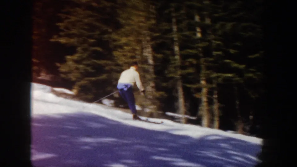 1961 Aspen frame showing the skier from behind on a sunlit snowy run bordered by evergreen trees.
