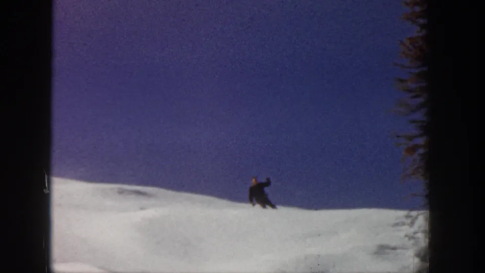 The Aspen skier descends a snow-covered slope near dark evergreen trees in grainy 1961 8mm footage.