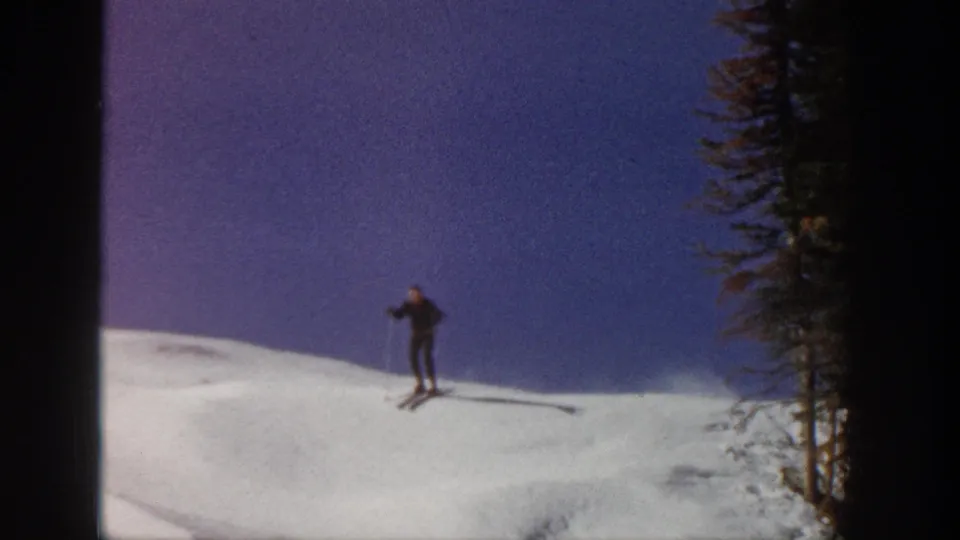 A skier pauses or slows on a snowy Aspen ridge, with evergreen trees and long shadows visible in the 1961 8mm frame.