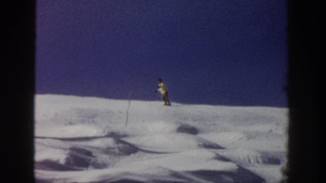 A small skier traverses a snowy Aspen ridge under blue sky, with a thin marker pole visible in the home-movie frame.