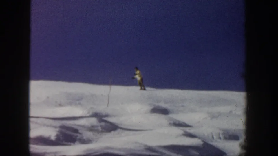 A small skier traverses a snowy Aspen ridge under blue sky, with a thin marker pole visible in the home-movie frame.