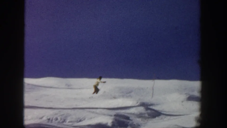 A distant skier leans into motion across the Aspen slope, with textured snow and dark 8mm frame borders visible.