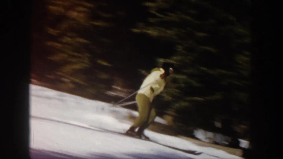 The skier passes nearer the camera beside evergreen trees, with yellow clothing, poles, and skis visible in warm 8mm color.