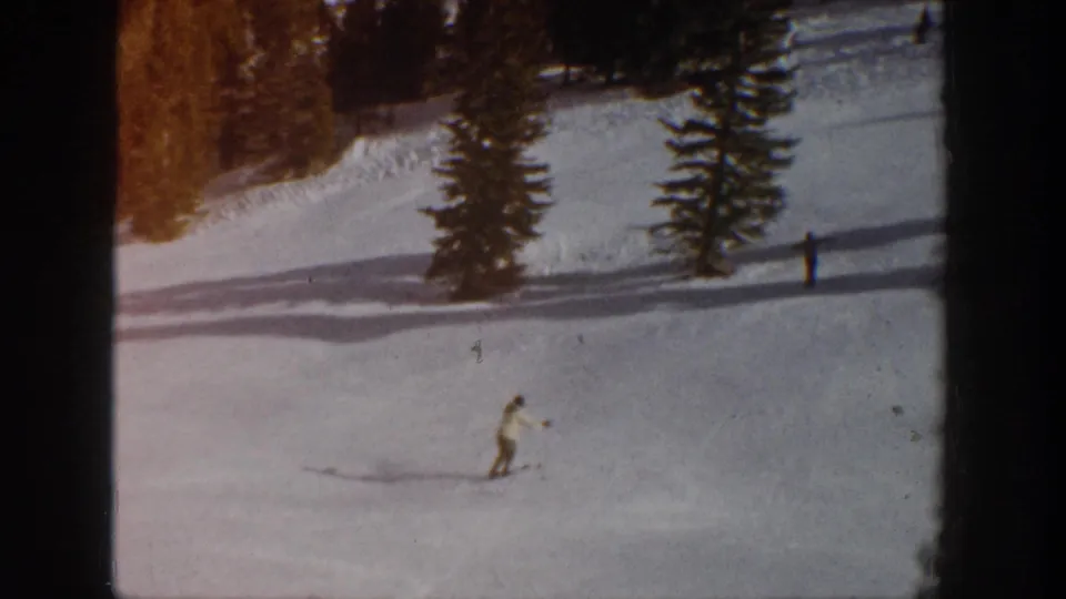 Wide 1961 Aspen home-movie view of a skier crossing open snow below evergreen trees and distant figures.
