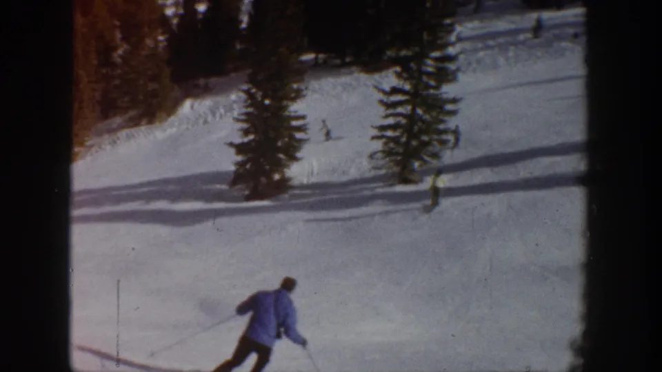 1961 Aspen ski-slope frame showing a skier in a blue jacket moving downhill past trees and distant skiers.