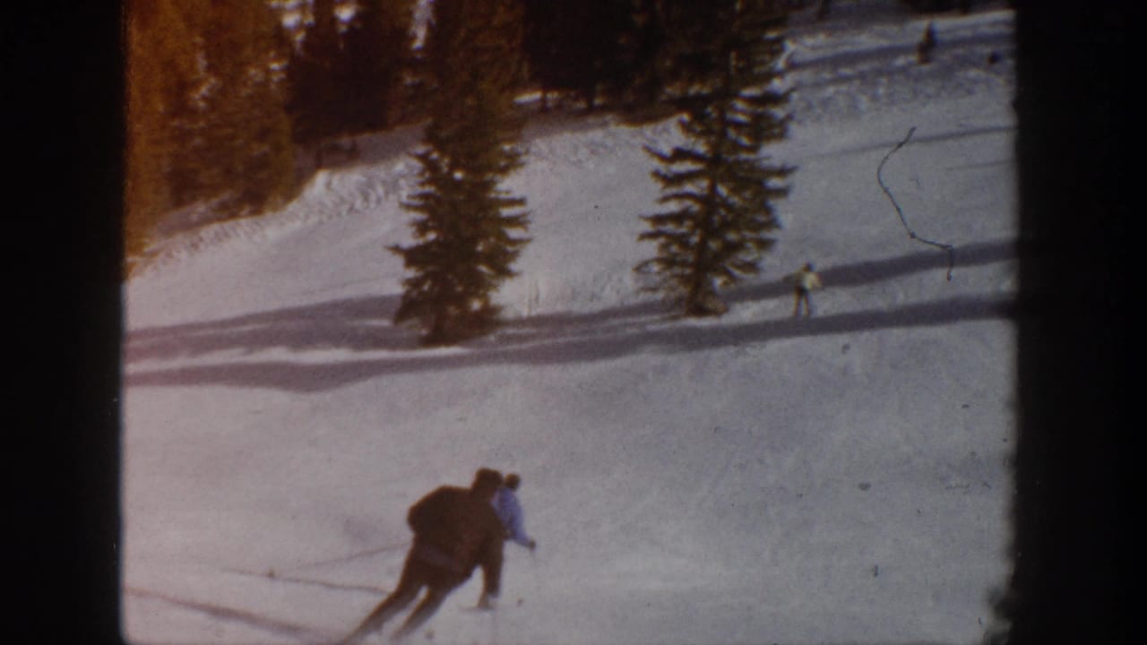 In the supplied 1961 Aspen, Colorado 8mm footage, two skiers move across a sunlit snowy slope with evergreen trees and long shadows behind them.