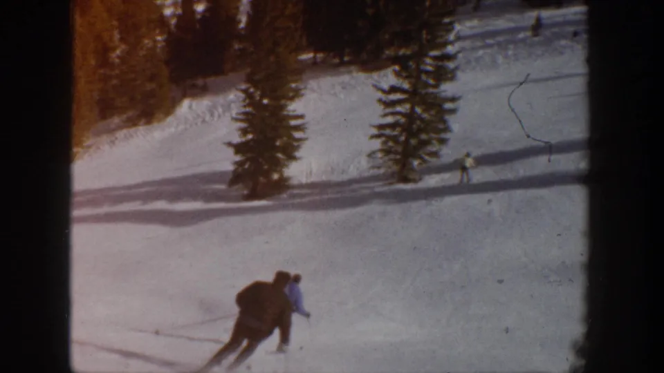 In the supplied 1961 Aspen, Colorado 8mm footage, two skiers move across a sunlit snowy slope with evergreen trees and long shadows behind them.