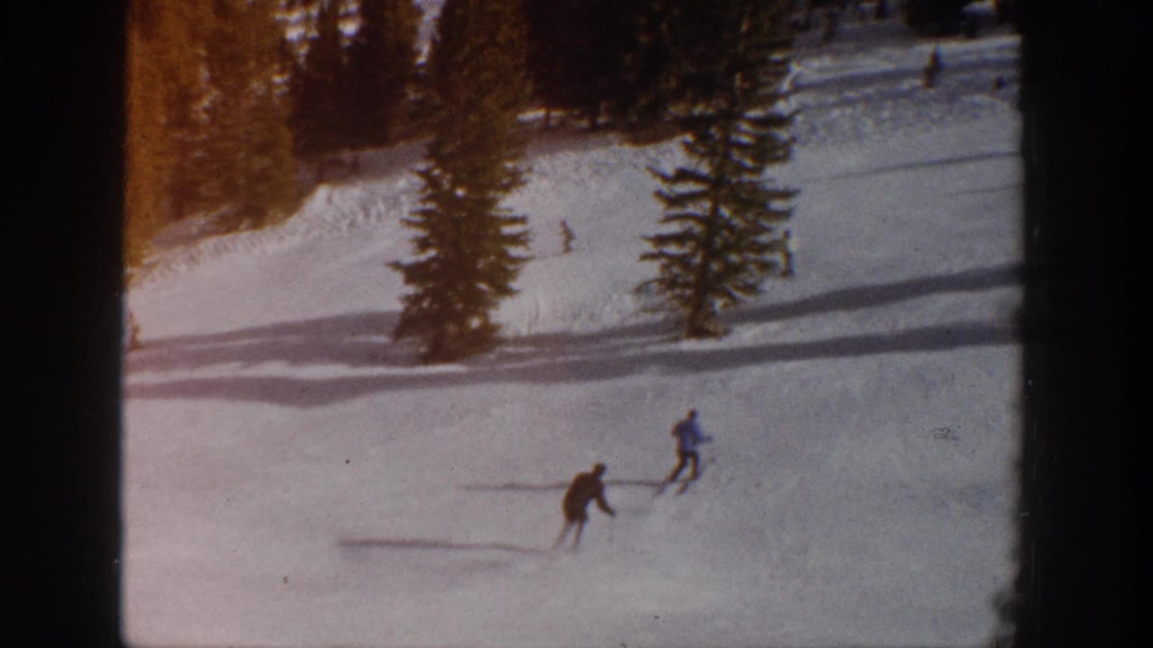 Two distant skiers in dark and blue winter clothing cross the Aspen snowfield, with pine trees, long shadows, and visible 8mm frame edges around the scene.