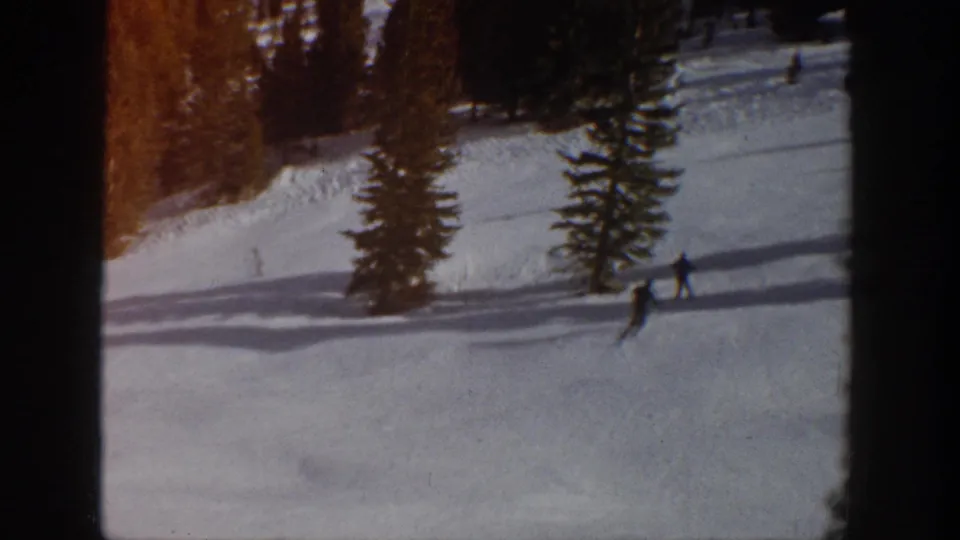 A distant skier crosses the lower part of a snowy Aspen slope while another figure appears farther uphill near evergreen trees and dark film-frame edges.