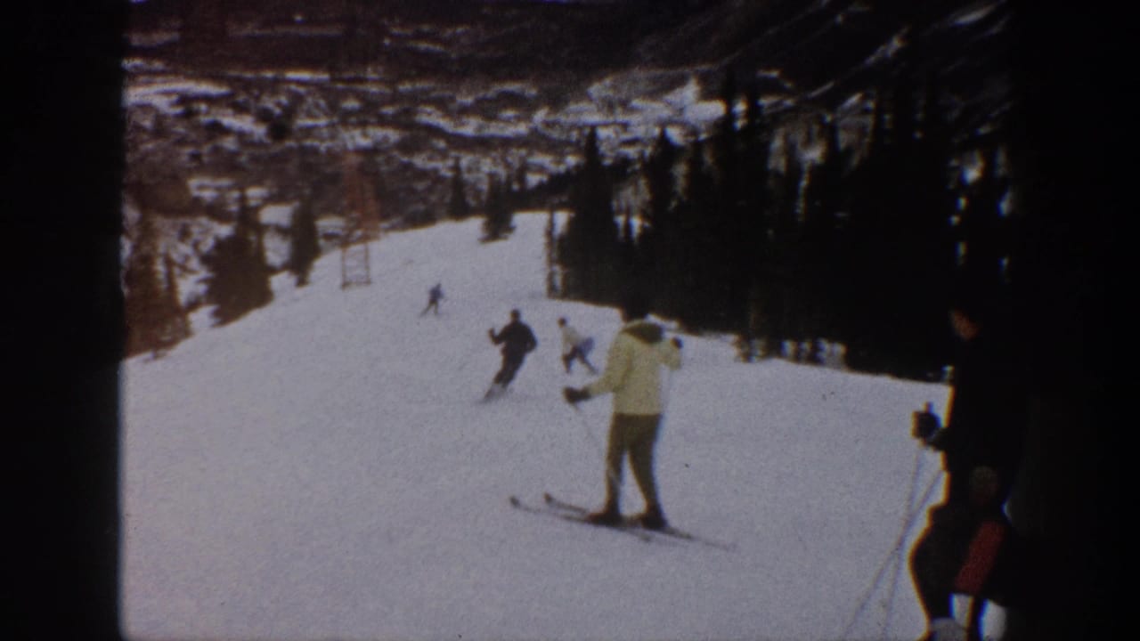 1961 Aspen home-movie frame showing several skiers on a snowy mountain slope with trees, lift structures, and a foreground skier in a light jacket.