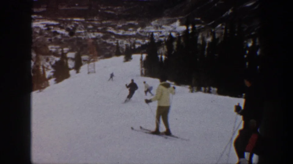 1961 Aspen home-movie frame showing several skiers on a snowy mountain slope with trees, lift structures, and a foreground skier in a light jacket.