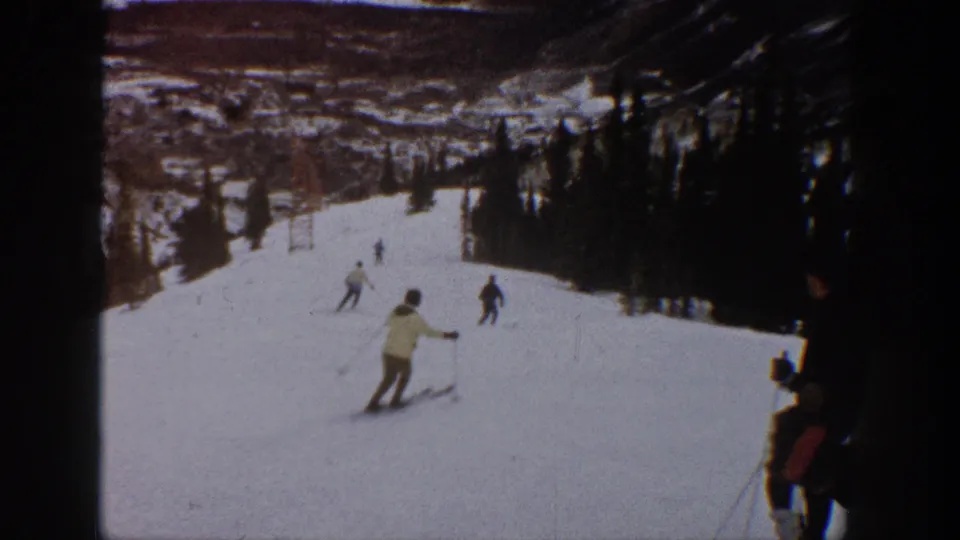 1961 Aspen ski-slope footage with a light-jacketed skier near center and other skiers spaced across the snowy run.