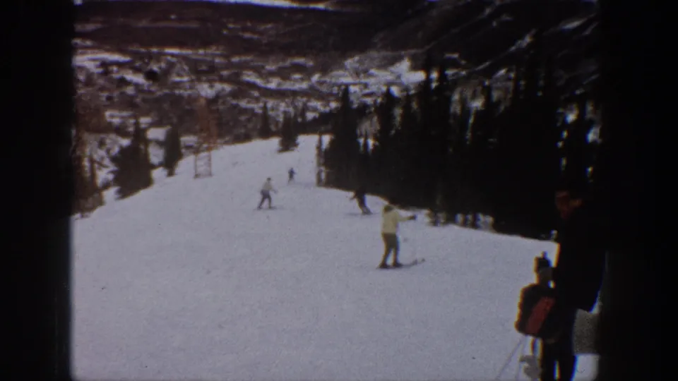 Home-movie view of an Aspen ski run in 1961, with a foreground skier near the right edge and distant figures on the snowy slope.