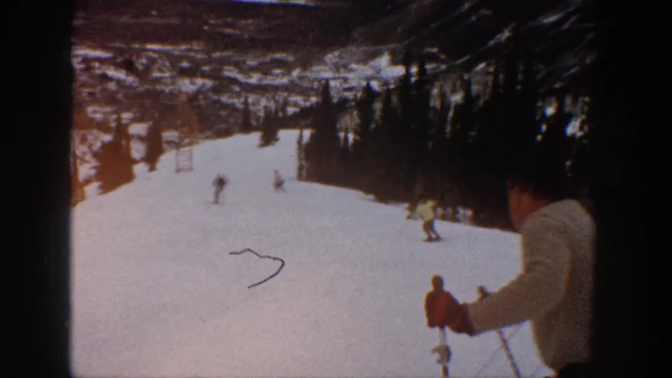 1961 Aspen vacation footage with a partially cropped foreground skier, distant skiers, snow, trees, and a visible mark or scratch on the film image.