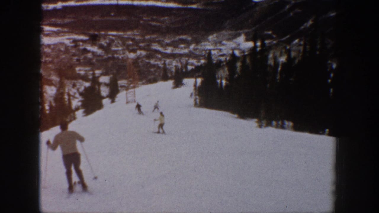 A skier in the foreground descends a broad snowy run with other skiers ahead.