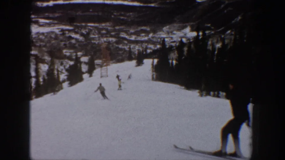 1961 Aspen home-movie frame with distant skiers on a snowy run and a partially cropped skier near the camera at right.