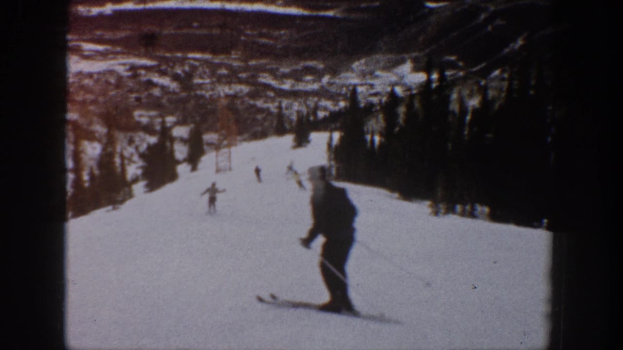 1961 Aspen home-movie frame showing distant skiers on a wide snowy run with trees and lift structures in the background.