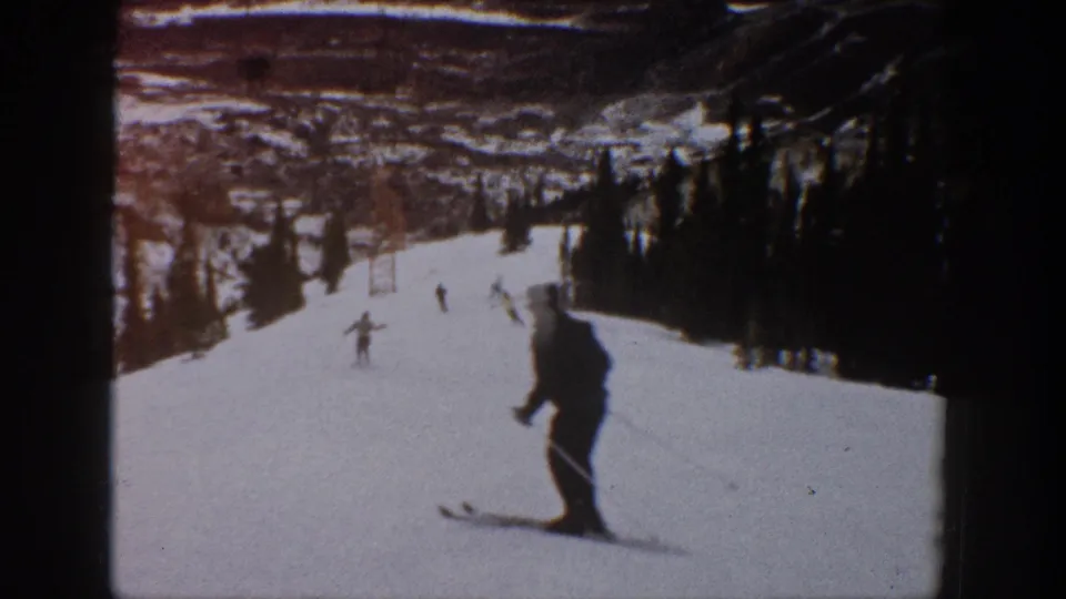 1961 Aspen home-movie frame showing distant skiers on a wide snowy run with trees and lift structures in the background.