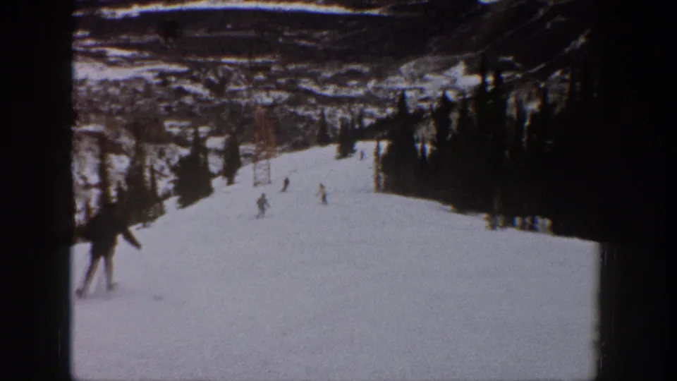 1961 Aspen ski footage frame with small figures descending a broad white slope, evergreen border, and lift towers visible beyond.