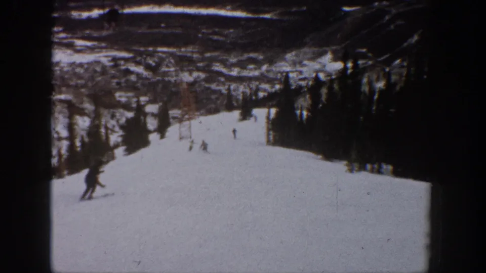 1961 Aspen home-movie frame showing a skier at lower left and smaller skiers farther down a snowy tree-lined run.