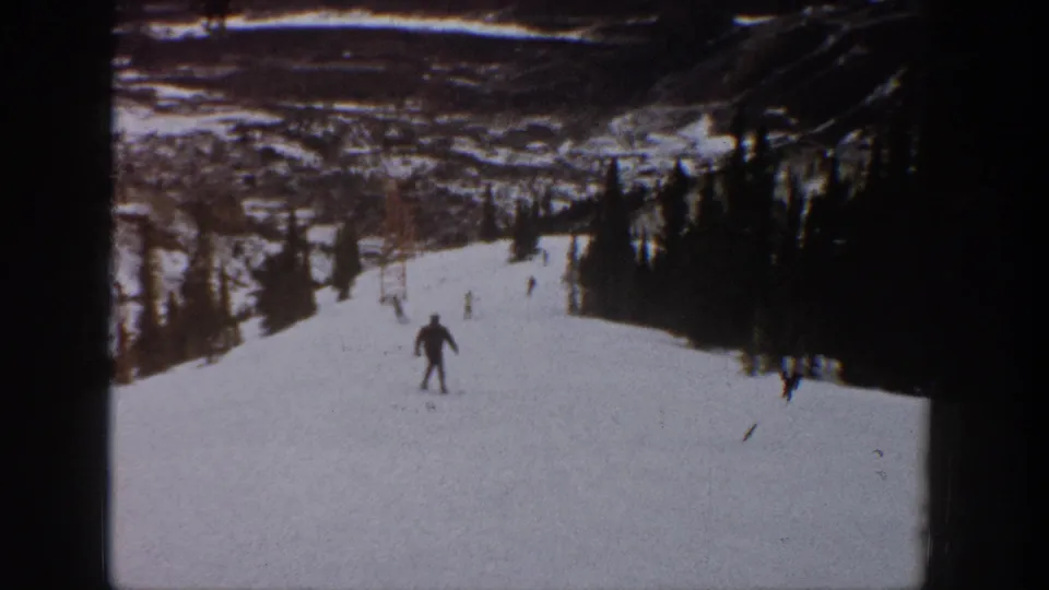 1961 Aspen frame showing a central skier on a broad snowfield, with trees, distant skiers, and lift structures beyond.