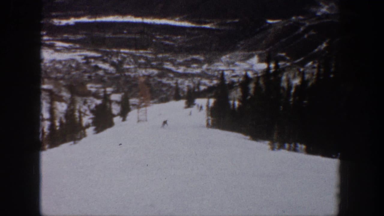 1961 Aspen home-movie frame showing small skiers on a wide snow-covered slope with trees, a lift tower, and distant mountain terrain.