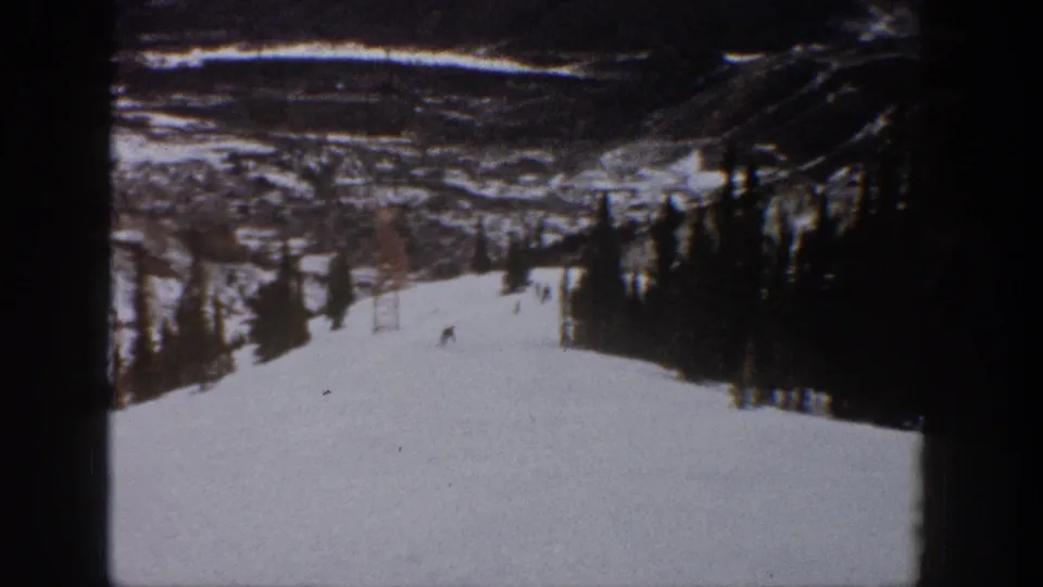 1961 Aspen home-movie frame showing small skiers on a wide snow-covered slope with trees, a lift tower, and distant mountain terrain.