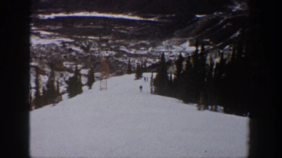 1961 Aspen skiing footage frame with a lone skier on a groomed snowy run, tree line at right, lift tower, and mountain backdrop.