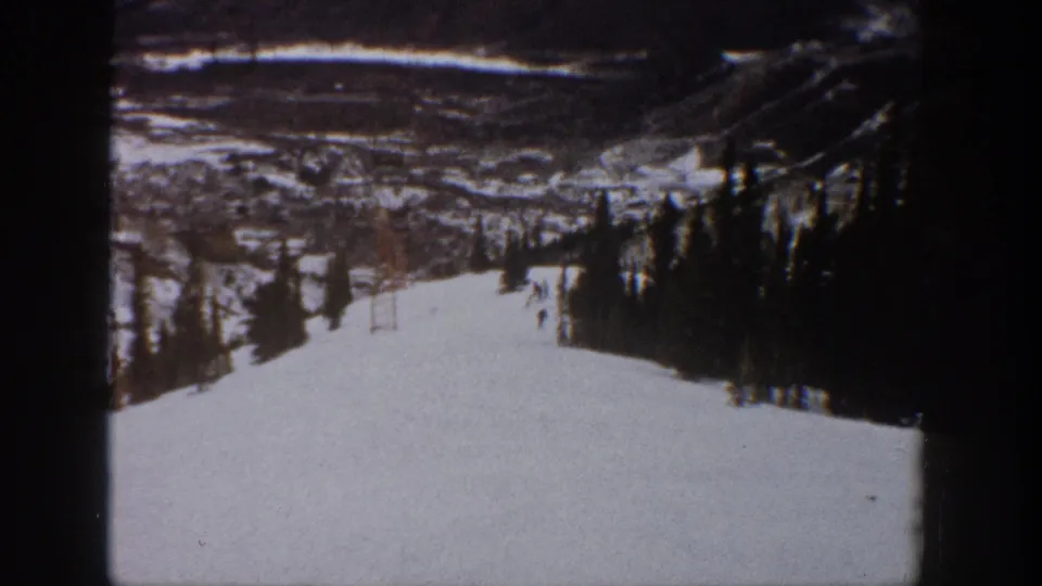 1961 Aspen home-movie still of skiers on a broad snow run, framed by dark trees and distant mountain slopes.