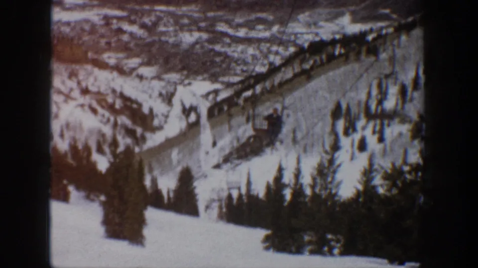 1961 Aspen, Colorado home-movie frame showing a snowy mountain ski slope with lift infrastructure, evergreen trees, and distant ridgelines.