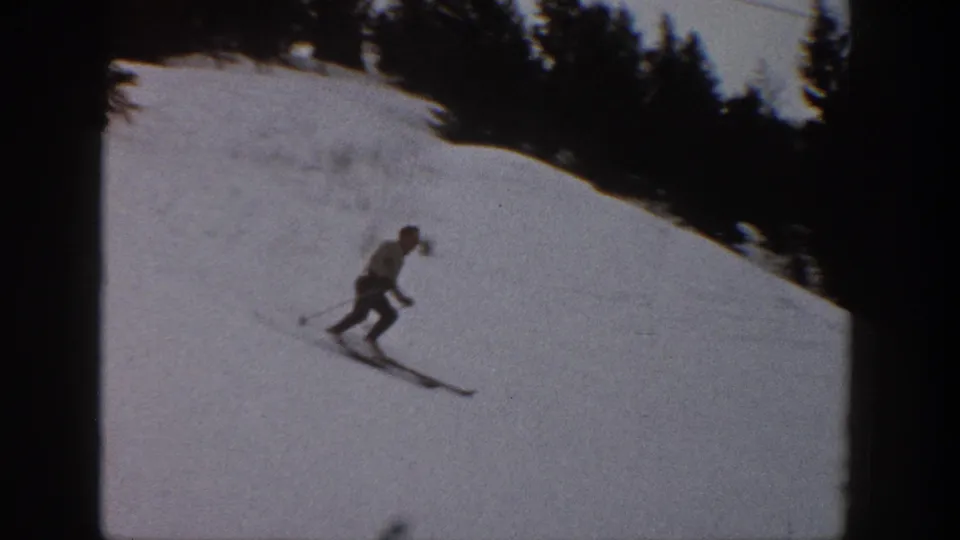 1961 Aspen, Colorado frame of a skier captured mid-descent on a snowy resort slope with evergreen trees behind.