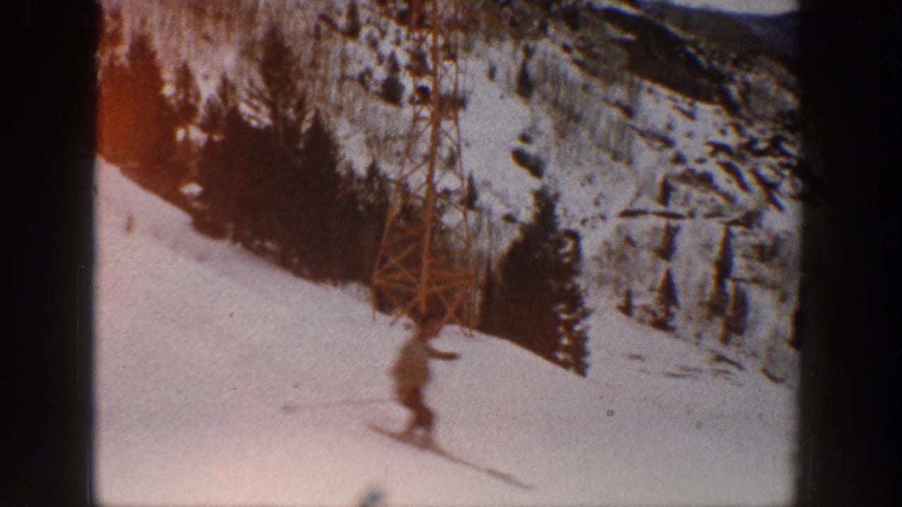 A skier on an Aspen snow slope passes near a tall tower, with evergreen trees and mountain terrain visible in the background.