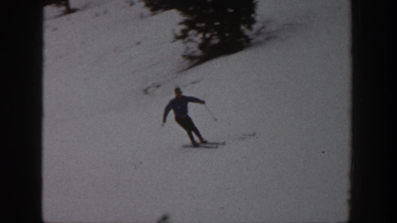 Adult skier in a blue jacket turns across the snowy Aspen slope, with trees visible high in the frame.