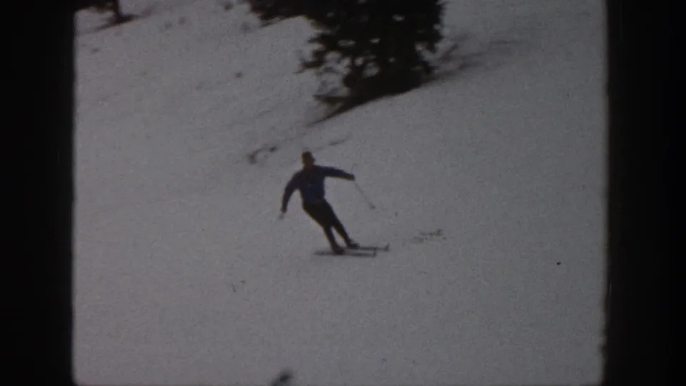 Adult skier in a blue jacket turns across the snowy Aspen slope, with trees visible high in the frame.
