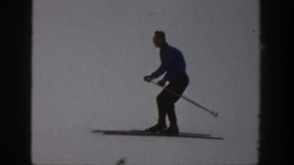 Skier in 1961 Aspen footage leans into a downhill glide, silhouetted against overexposed snow.