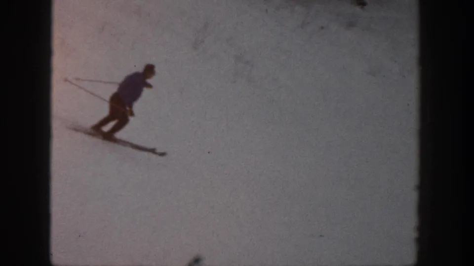 A distant skier in dark clothing leans into a turn on the Aspen snow, recorded with soft 8mm home-movie texture.