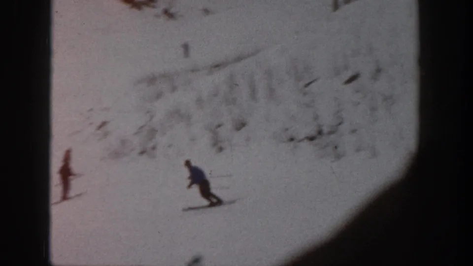 Distant skiers move downhill on a snowy Aspen slope, with rocky patches visible along the upper edge.
