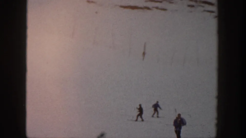Several small figures in winter clothing stand or ski across a broad snowy Aspen slope in faded 8mm color.