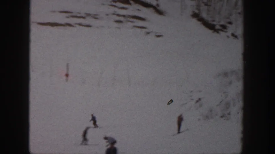 1961 Aspen home-movie frame showing small skiers crossing a snow-covered slope, with film grain, scratches, and dark frame edges.