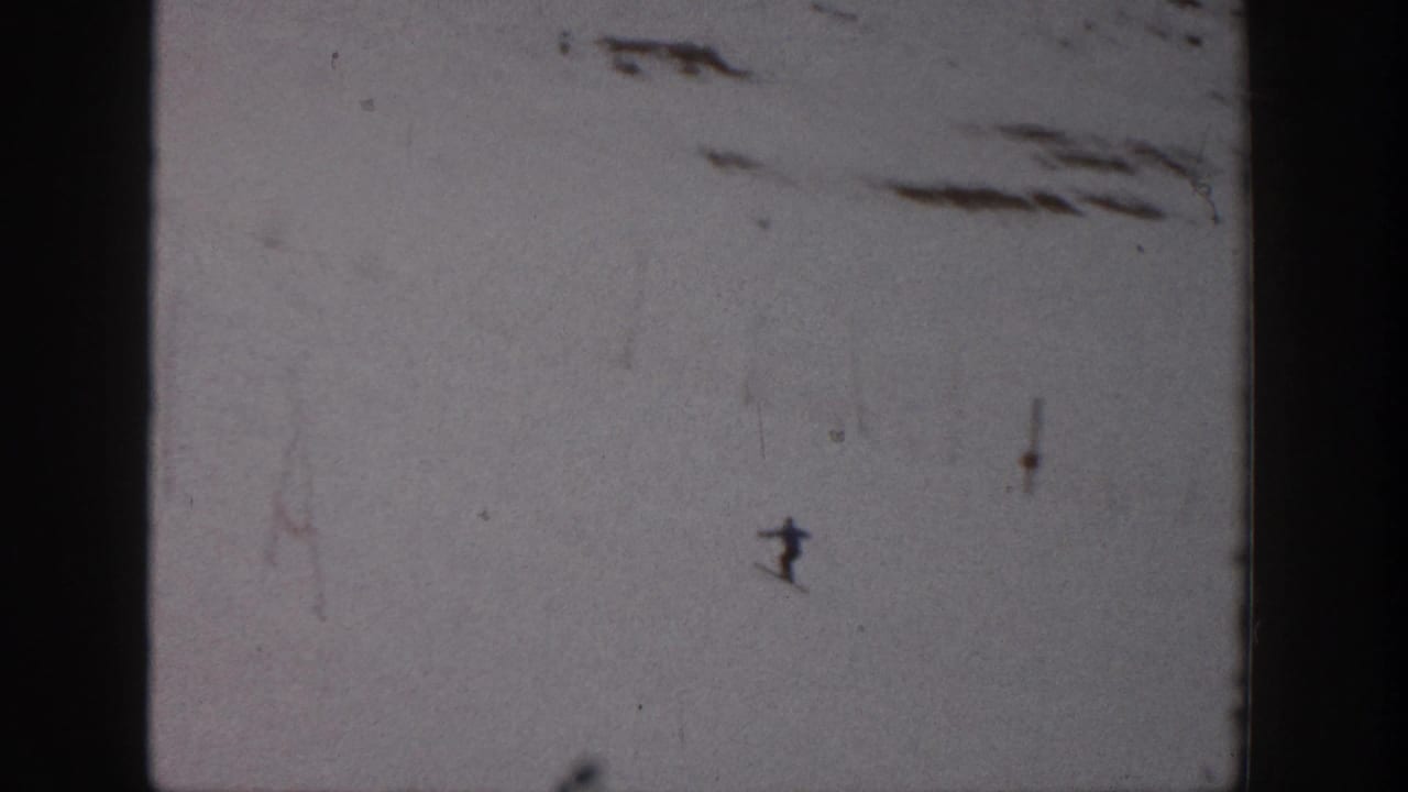 A small skier descends a wide snowy slope near sparse dark patches.