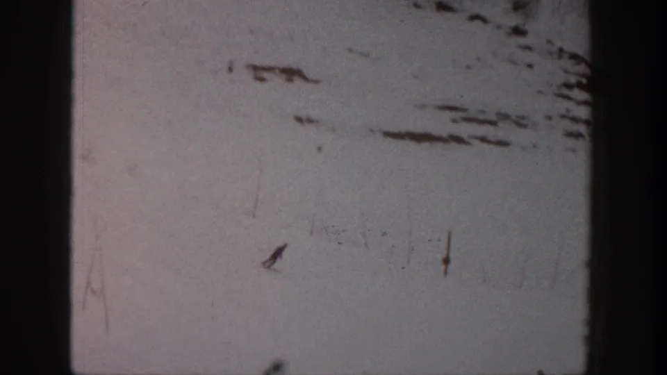 1961 Aspen ski footage shows a small skier angled across the snow, with scratches, dust, and uneven home-movie exposure.