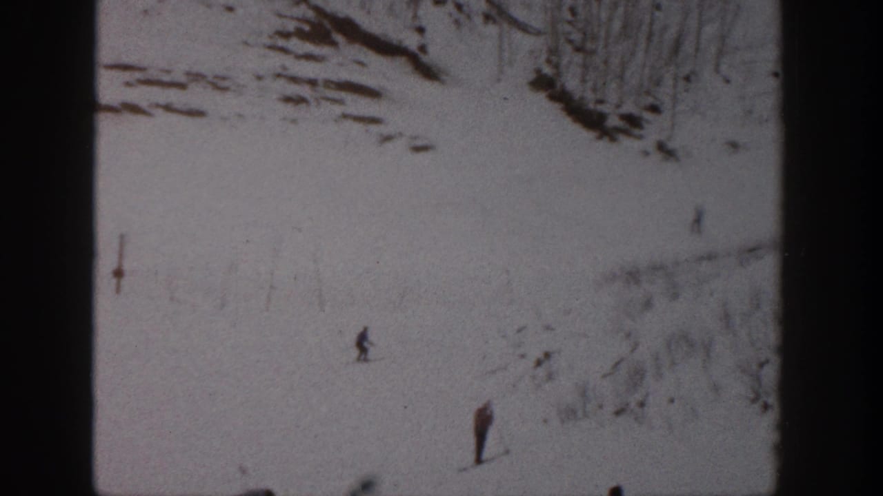 1961 Aspen home-movie frame showing small skiers spaced across a snow-covered slope with rocky terrain above.