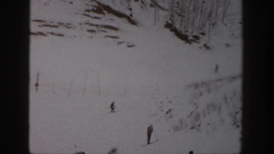 1961 Aspen home-movie frame showing small skiers spaced across a snow-covered slope with rocky terrain above.