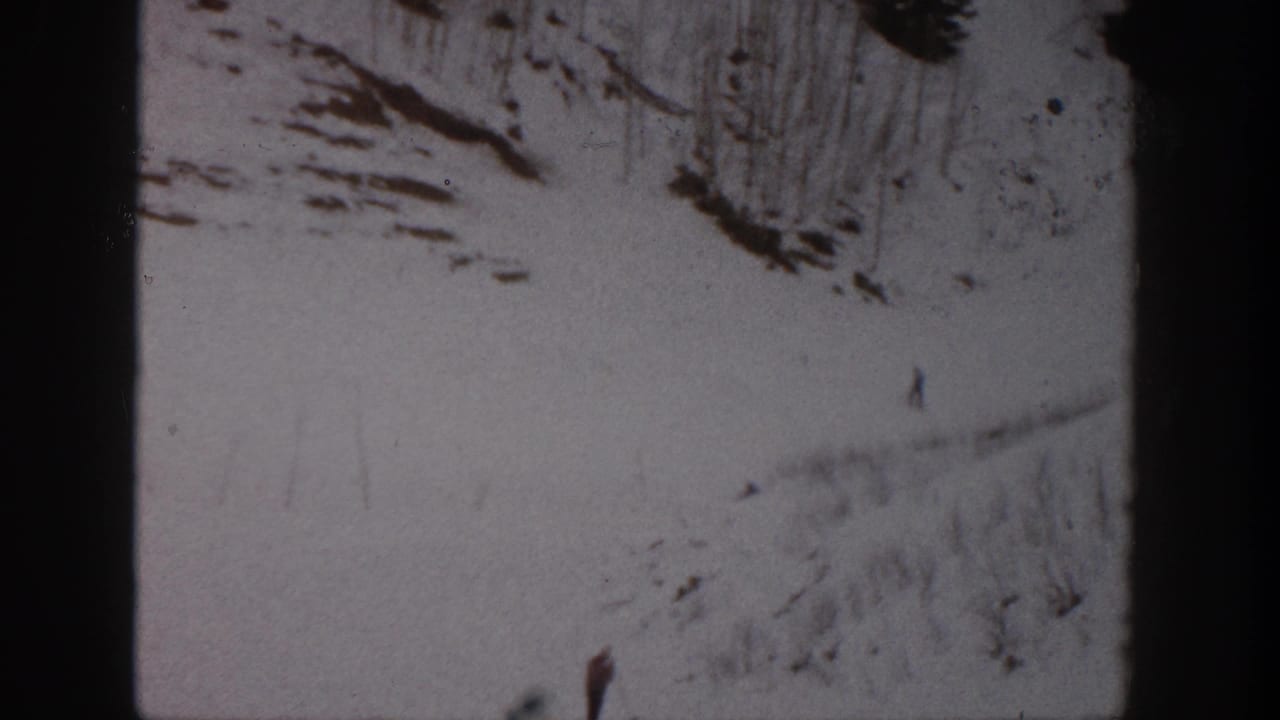 Snowy hillside with several tiny skiers and rocky patches near the top.
